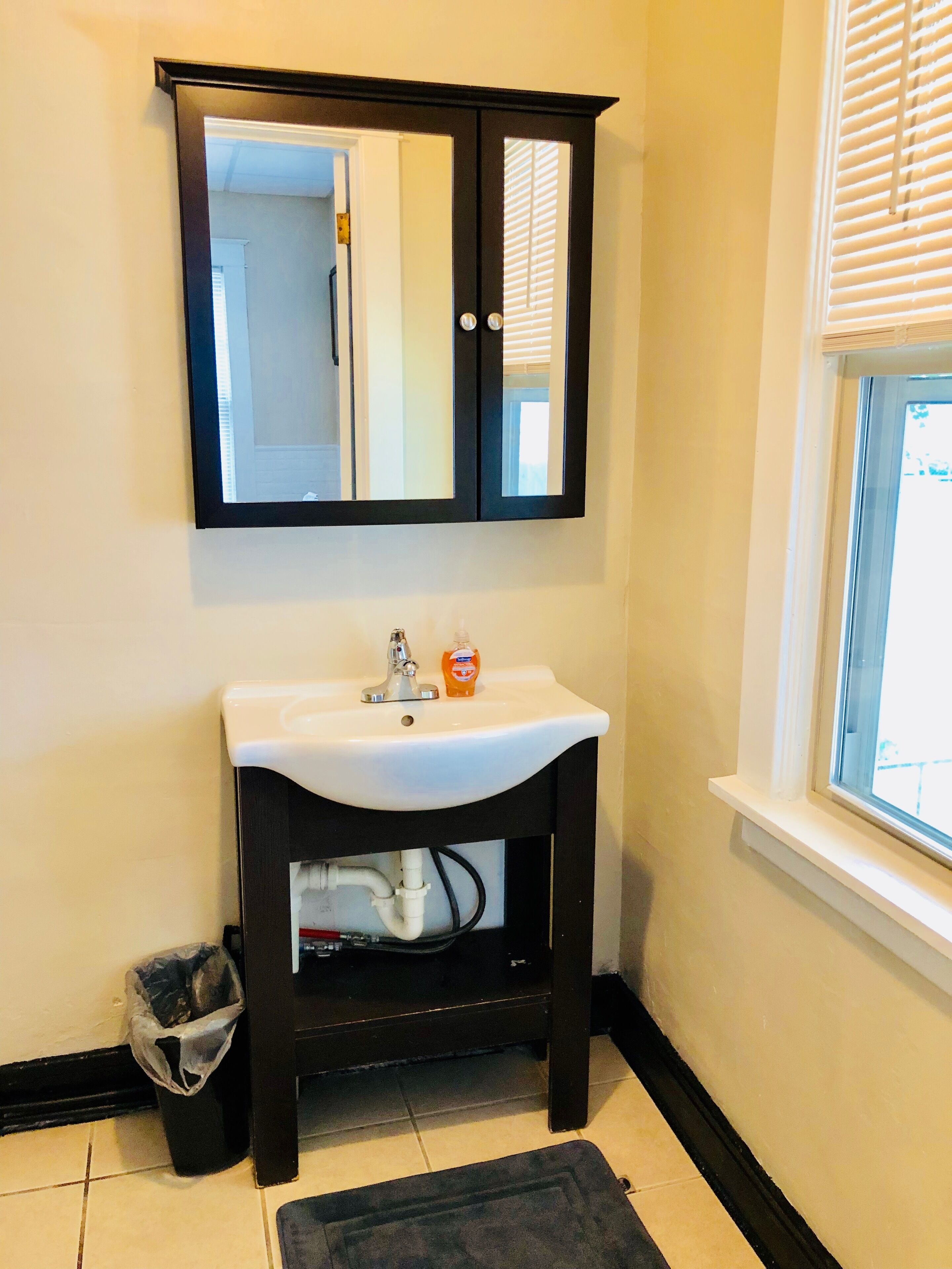 Un baño con un lavamanos blanco, un mueble de madera oscura, un espejo y una ventana con persianas.
