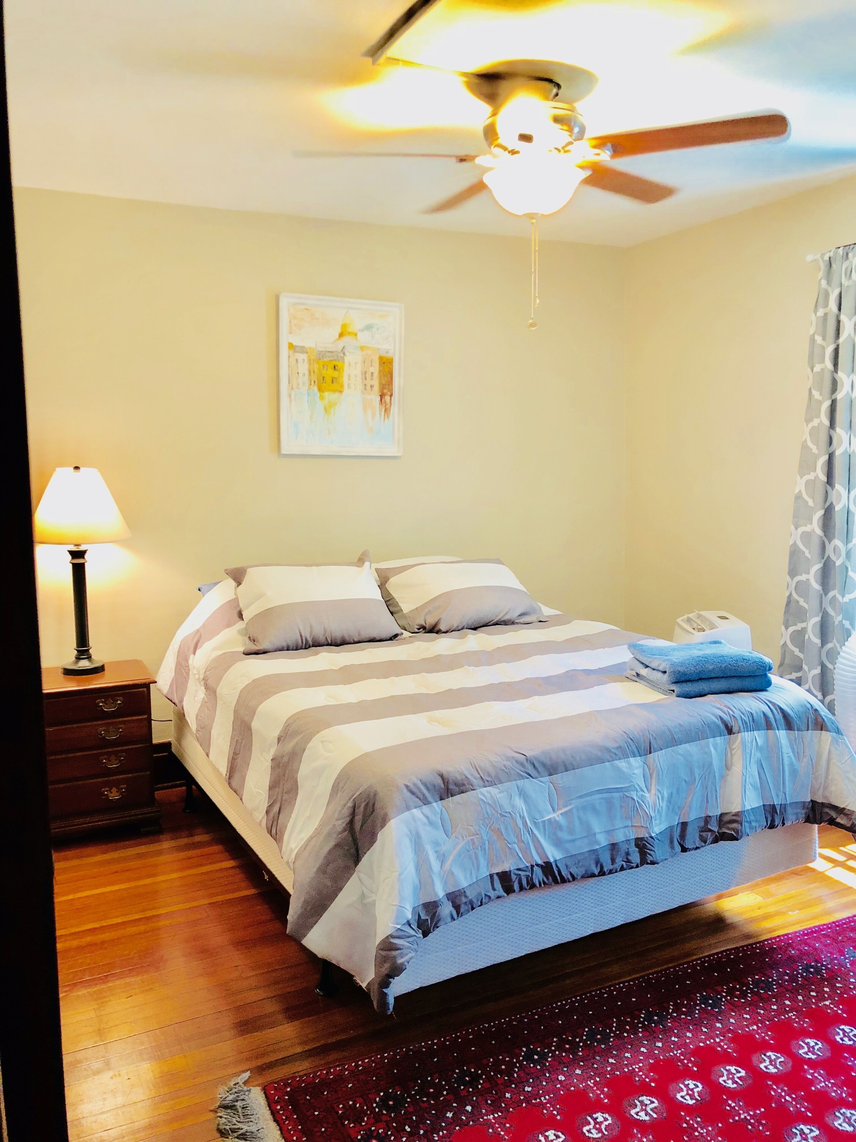 A bedroom with a bed, a nightstand, a lamp, a ceiling fan, and a painting on the wall.