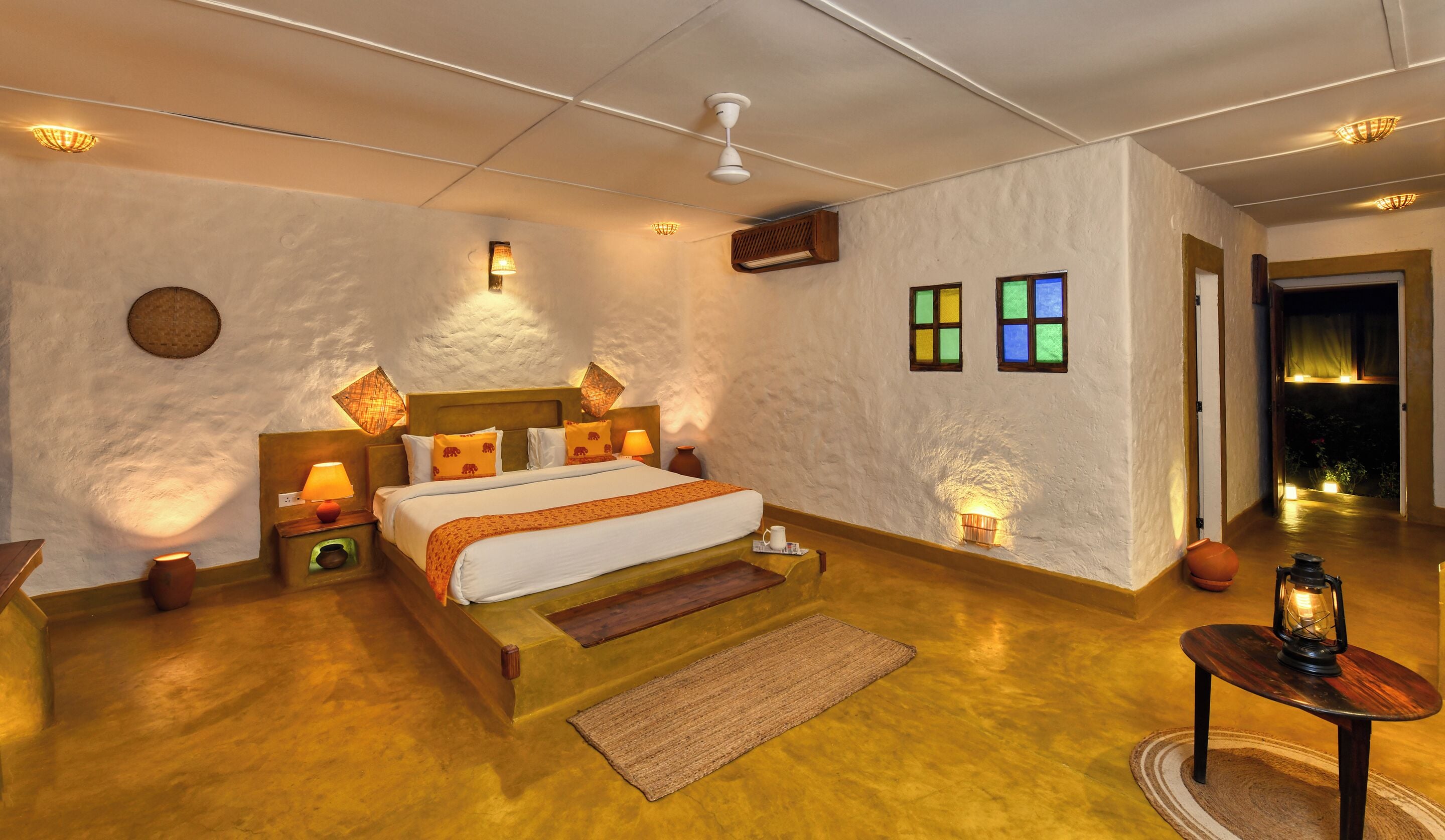 Un dormitorio con una cama grande, lámparas de noche, una mesita con un farol y un ventanal con vidrios de colores.