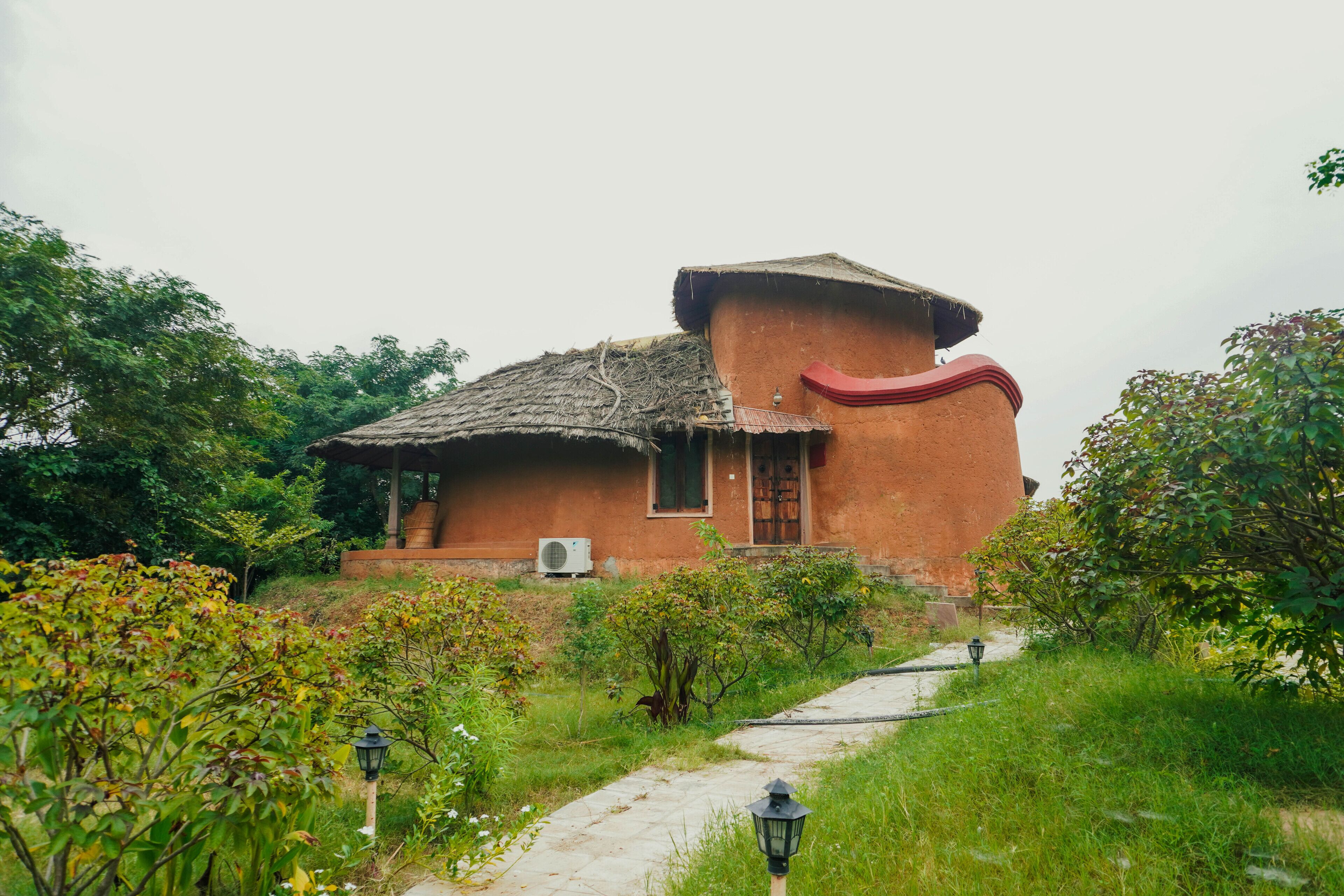 A rustic house with a thatched roof, surrounded by lush greenery and a paved pathway.