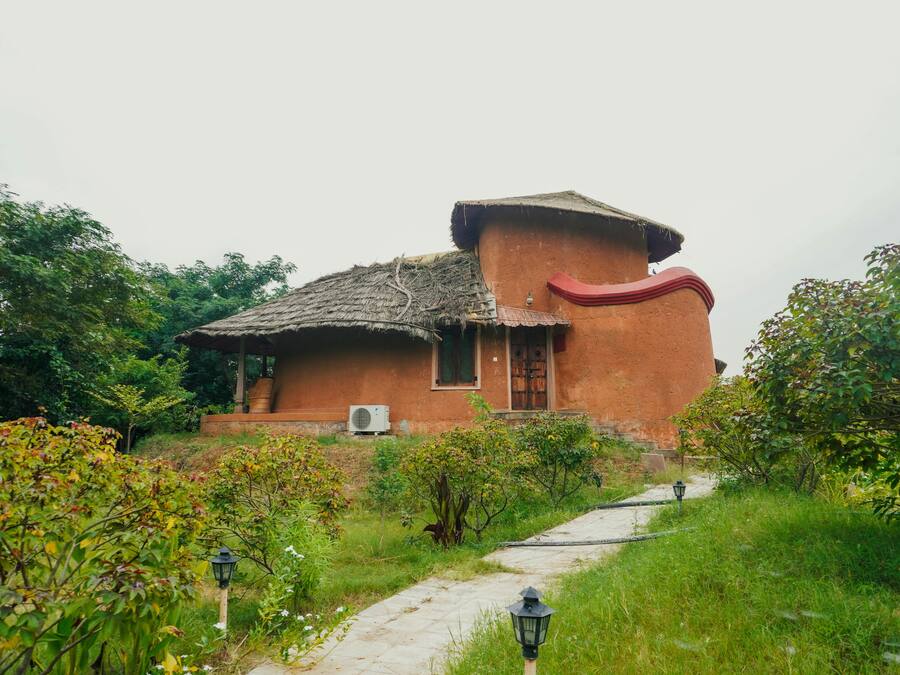 A rustic house with a thatched roof, surrounded by lush greenery and a paved pathway.
