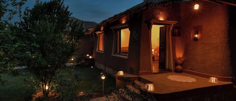A rustic building with a thatched roof, illuminated windows, and a small porch area.