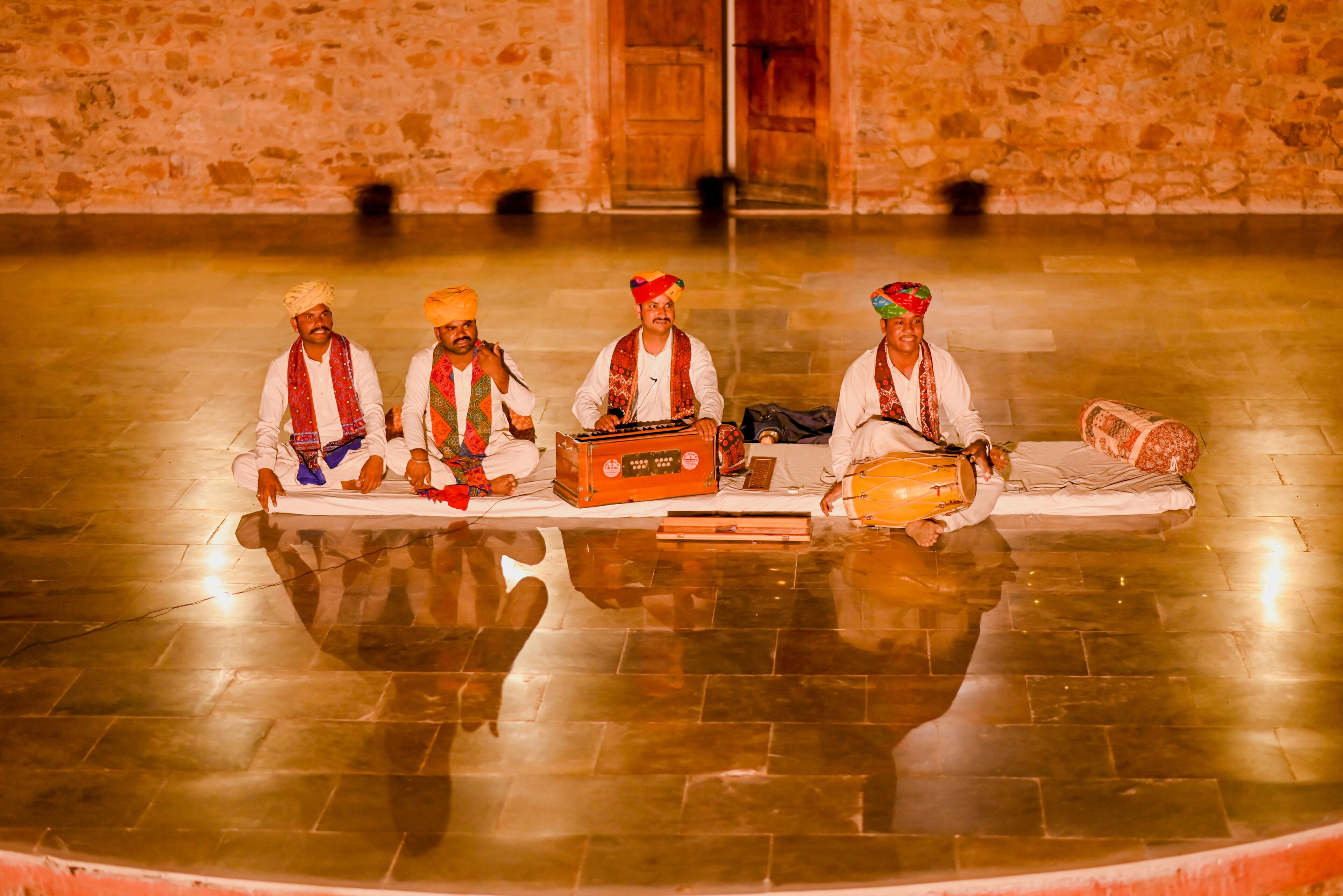 Four individuals seated on a reflective floor, playing traditional instruments in a room with stone walls and a wooden door.