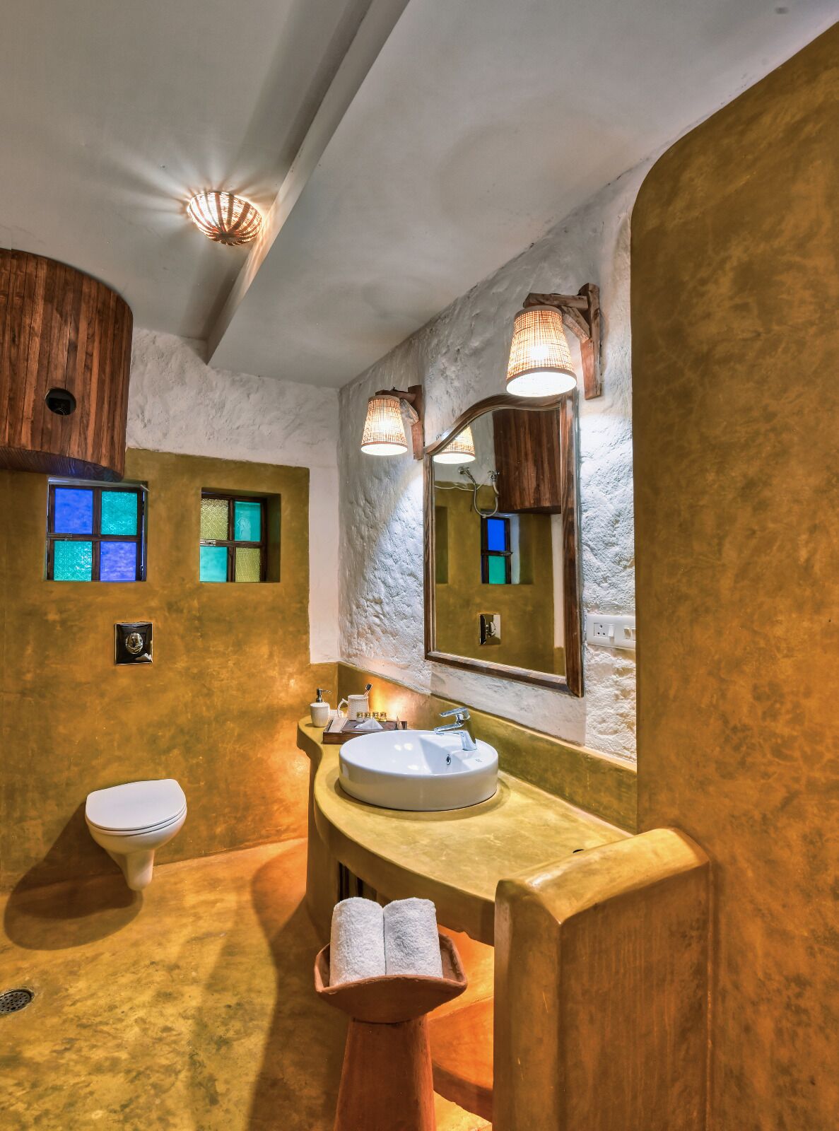 A bathroom with a round sink, a toilet, and a mirror. There are two windows with stained glass, and a wooden shelf with toiletries.