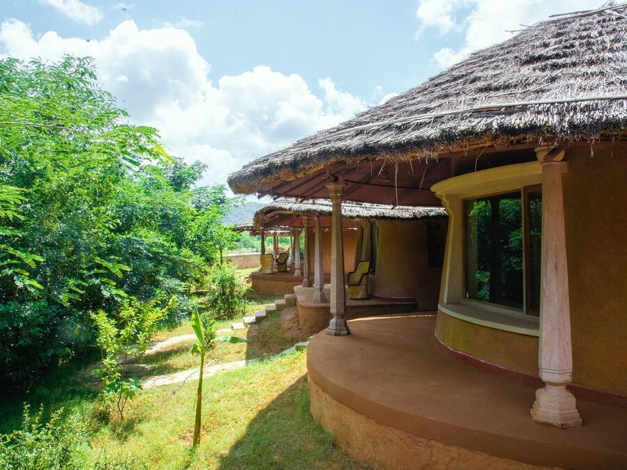 A traditional thatched-roof building with a covered veranda, surrounded by lush greenery and a clear sky.