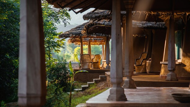 A covered outdoor seating area with stone columns and chairs, surrounded by greenery and steps leading up to a structure with a thatched roof. - The Earth At Ranthambore (Sawai Madhopur)