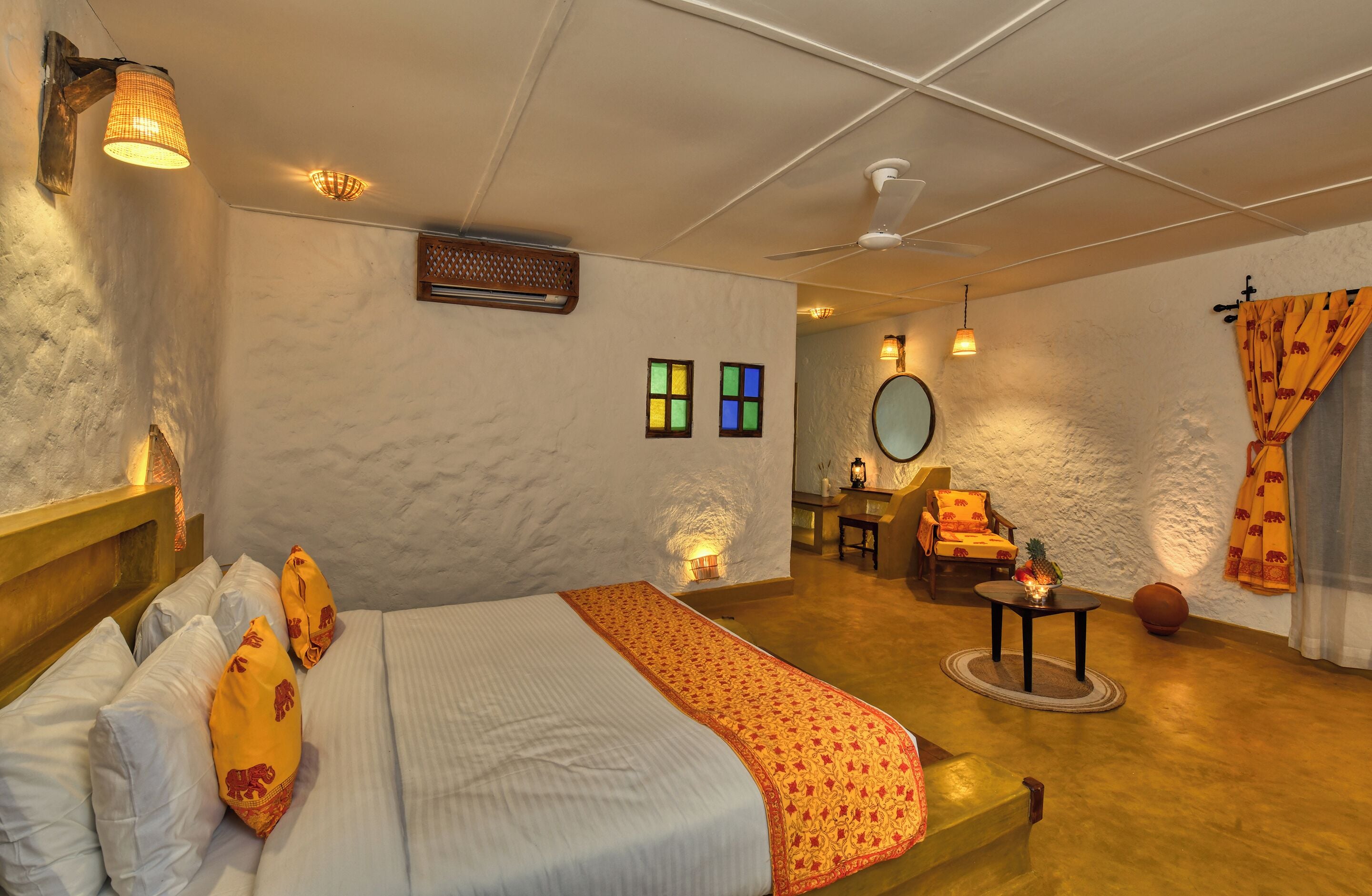 Una habitación con cama, una mesita de noche con lámpara, una silla y un espejo redondo en la pared.