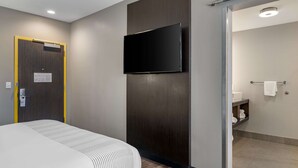 A hotel room with a bed, a mounted television, a door with a sign, and a bathroom visible through an open door.