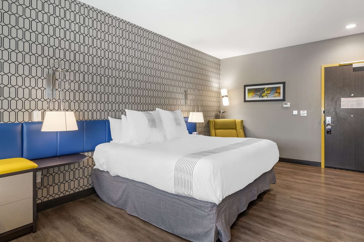 Hotellihuone, jossa on kuviollinen seinä, valkoisilla lakanoiden peitetty sänky, sininen pääty ja keltainen nojatuoli sekä puulattia.