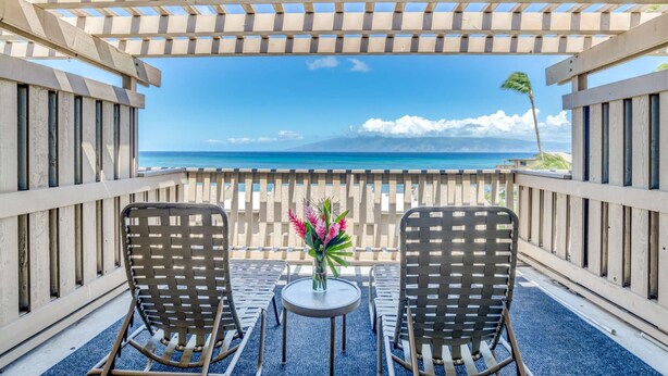 A balcony with two wicker chairs and a small table, overlooking a beach with clear skies and a mountain in the distance. - Kahana Sunset E10:  Summer and Fall Savings!  Free Activities! (Kahana)