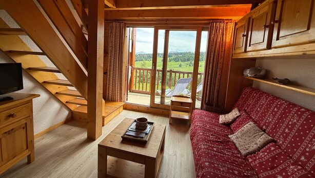 A wooden cabin interior with a sofa, a table, a TV, and a balcony with a view of the countryside. - South Apartment, 8 pers, 44m², panoramic slopes (Les Saisies)