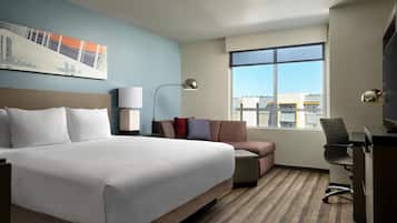 A hotel room with a large bed, a desk, a chair, a lamp, and a window with a city view.