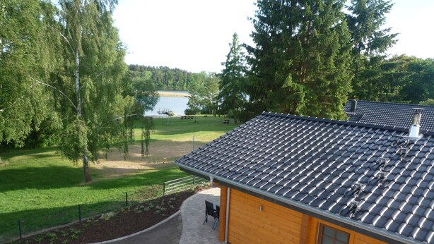 A wooden cabin with a tiled roof, surrounded by a paved driveway and a fenced garden area. - Ferienhaus am See in Fleeth (Mirow)