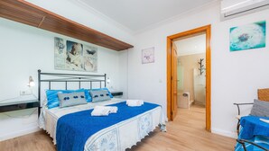 A bedroom with a bed, a bedside table, a chair, and a door leading to a bathroom. - Holiday Home ’Casa Paqui Alcudia’ with Wi-Fi, Garden, Terraces & Pool (Puerto de Alcudia)