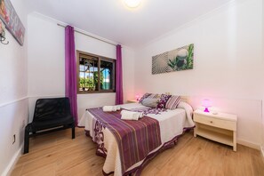 Una camera da letto con un letto, un comodino, una sedia, una finestra con tende e un quadro appeso al muro.
