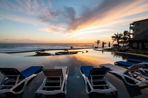 An infinity pool with lounge chairs, palm trees, and a view of the ocean at sunset.