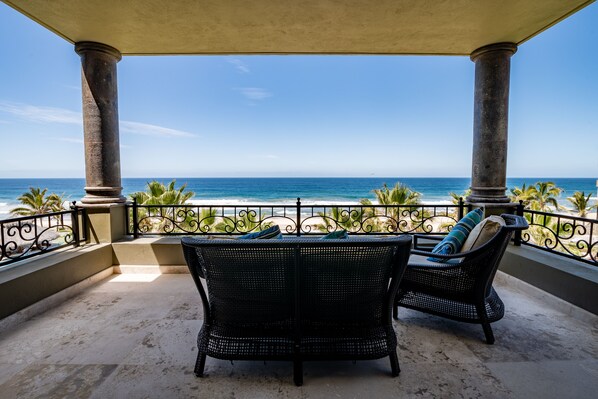 A balcony with two wicker chairs, a view of the ocean, and a railing with palm trees.