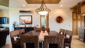 A dining area with a wooden table, wicker chairs, a central light fixture, a painting on the wall, and a kitchen in the background.