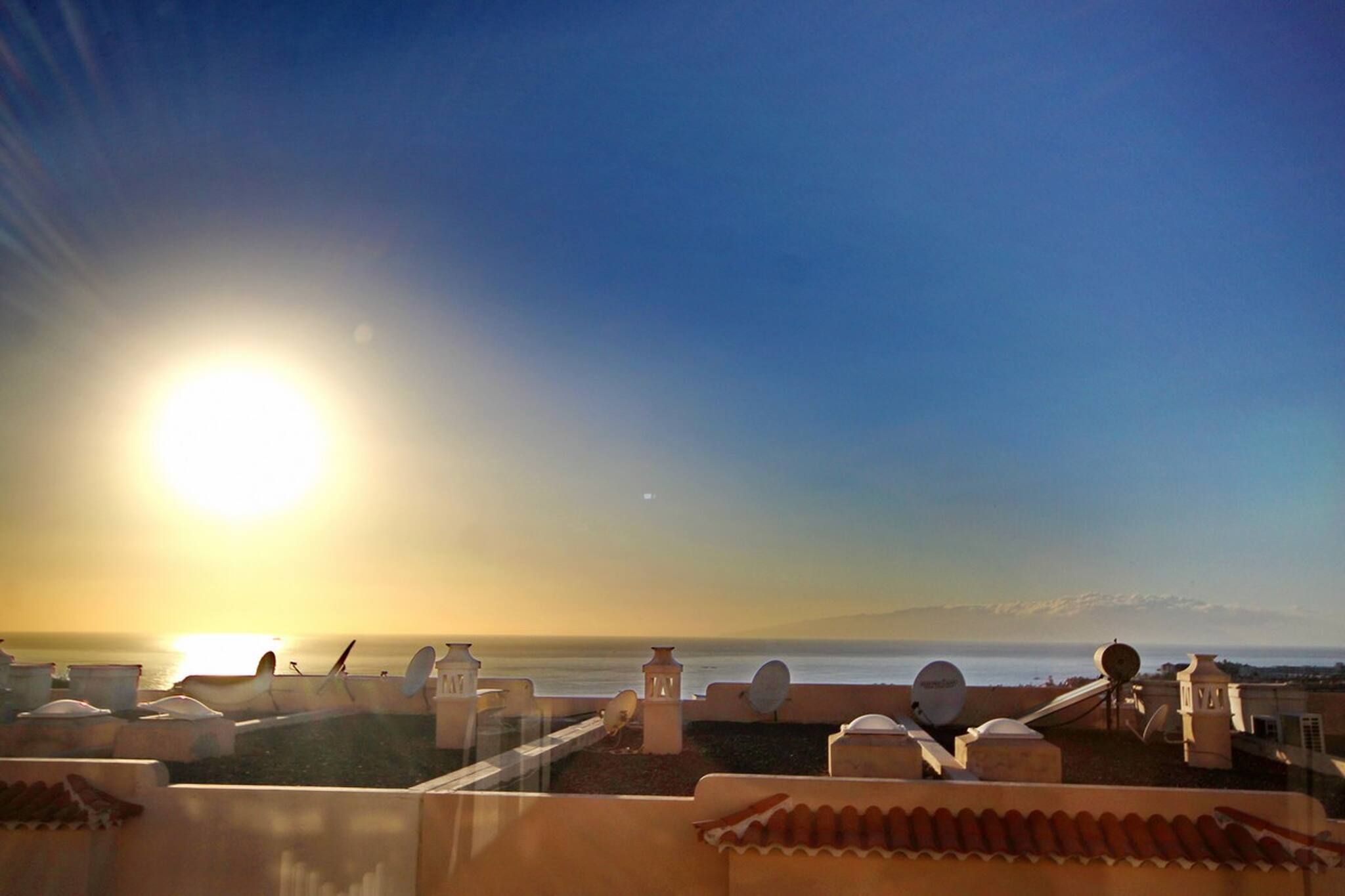 Una terraza con vista al océano al atardecer, con mobiliario exterior y antenas parabólicas.