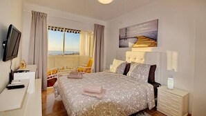 A hotel room with a bed, a desk, a chair, a TV, a painting, and a window with curtains.