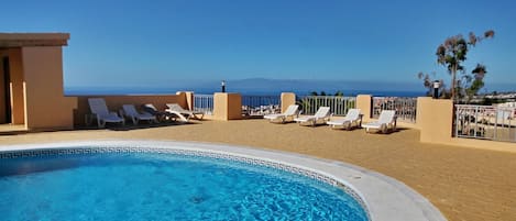 A pool with clear blue water, surrounded by lounge chairs and a view of the sea.