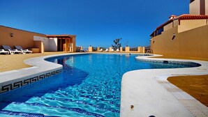 A swimming pool with a clear blue water feature, surrounded by lounge chairs and a building with a tiled roof.
