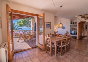A spacious living area with a wooden dining table and chairs, a large window with a view of the sea, and a staircase leading to an upper floor.