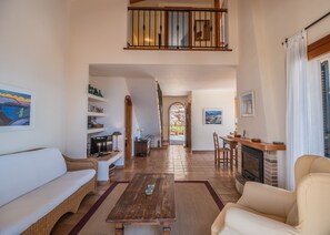 A spacious living area with a high ceiling, a wooden staircase, a fireplace, and a view through an arched doorway.