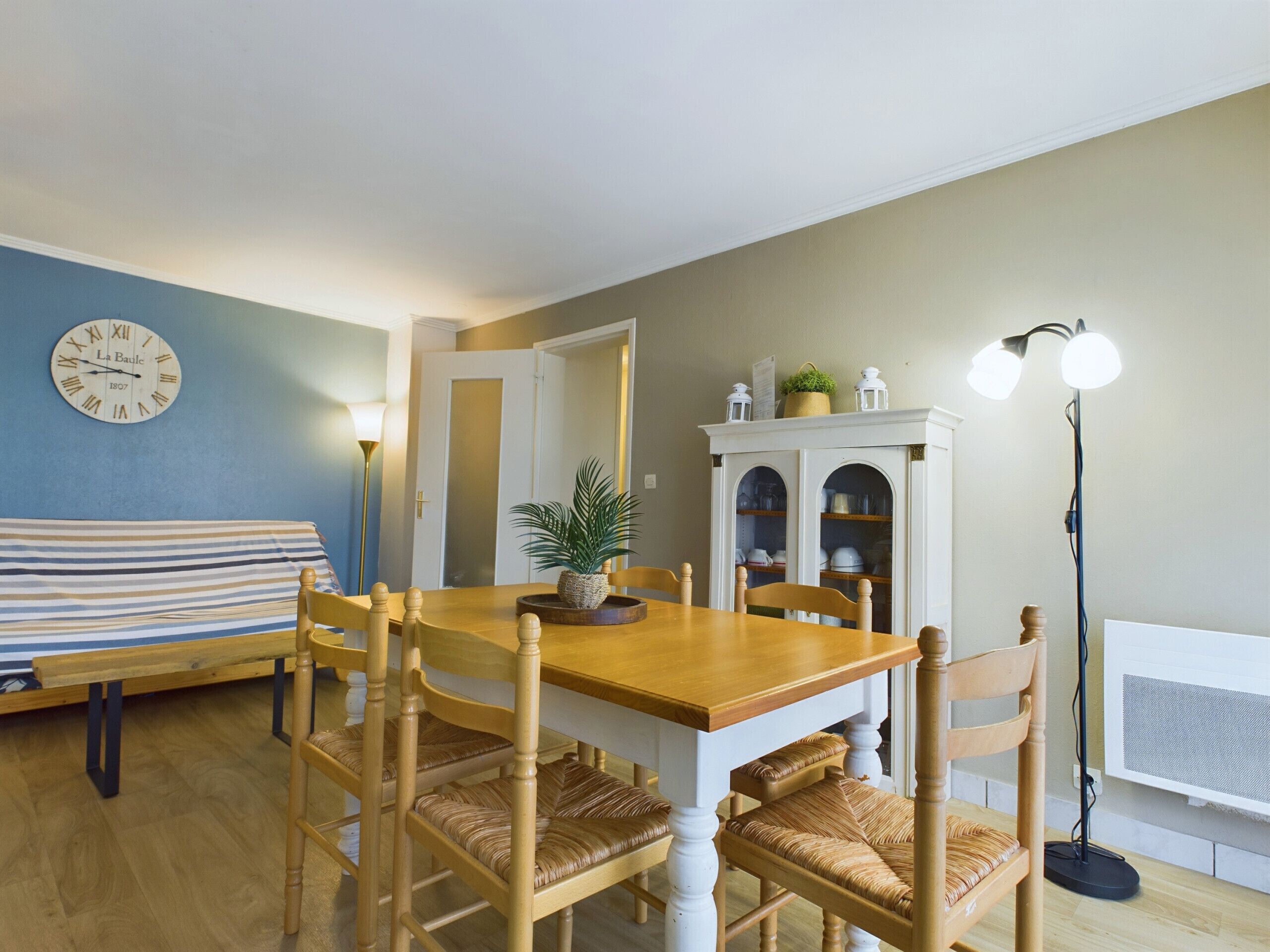 Een eethoek met een houten tafel en stoelen, een klok aan de muur, een lamp en een kast met servies.