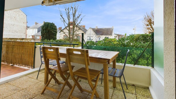 Een buitenterras met een houten tafel en stoelen, een overdekt gedeelte en een uitzicht op huizen in de verte.