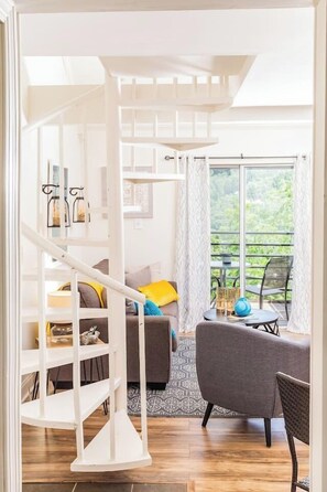 Stunning spiral staircase leads to a cozy seating area with a sofa and wicker chair, offering a view of lush greenery.