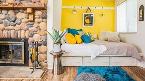 Une pièce avec une cheminée en pierre, un lit agrémenté de coussins colorés, un mur jaune et un tapis bleu.
