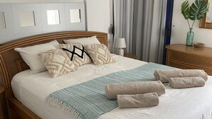Una cama bien hecha con edredón blanco, varios almohadones, una manta de color azul y blanco, y toallas enrolladas con texto.