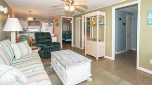 A living room with a sofa, a chair, a white chest of drawers, a ceiling fan, and a kitchen in the background.