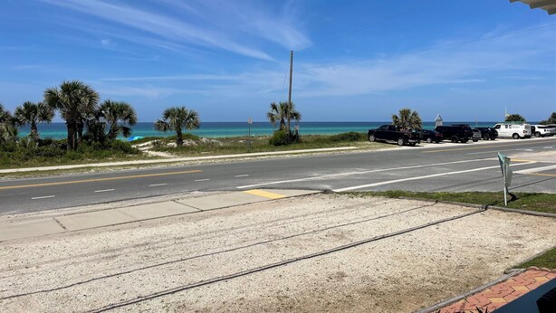 A coastal road with a clear view of the ocean, palm trees, and parked vehicles.