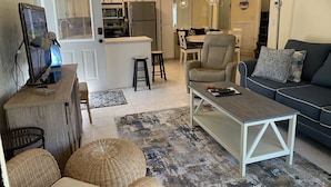 A living room with a sofa, coffee table, and ottomans. There is a kitchen area with a refrigerator, microwave, and bar stools. - Happy Hours I New Smyrna Poolside at the Beach 2bd/2ba +loft (New Smyrna Beach)