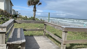 A wooden deck with a bench, overlooking a beach with waves and a palm tree. - Happy Hours I New Smyrna Poolside at the Beach 2bd/2ba +loft (New Smyrna Beach)