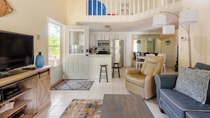 A spacious living room with a television, sofa, and coffee table. A kitchen is visible in the background with white cabinets and a dining area. - Happy Hours I New Smyrna Poolside at the Beach 2bd/2ba +loft (New Smyrna Beach)