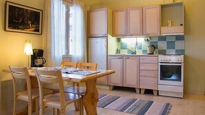 A compact kitchen with wooden cabinets, a dining table with chairs, a refrigerator, a stove, and a window with curtains.