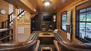 A cozy cabin interior with a wooden ceiling, a dining area with a table and chairs, a fireplace, and a large window offering a view of the outdoors.