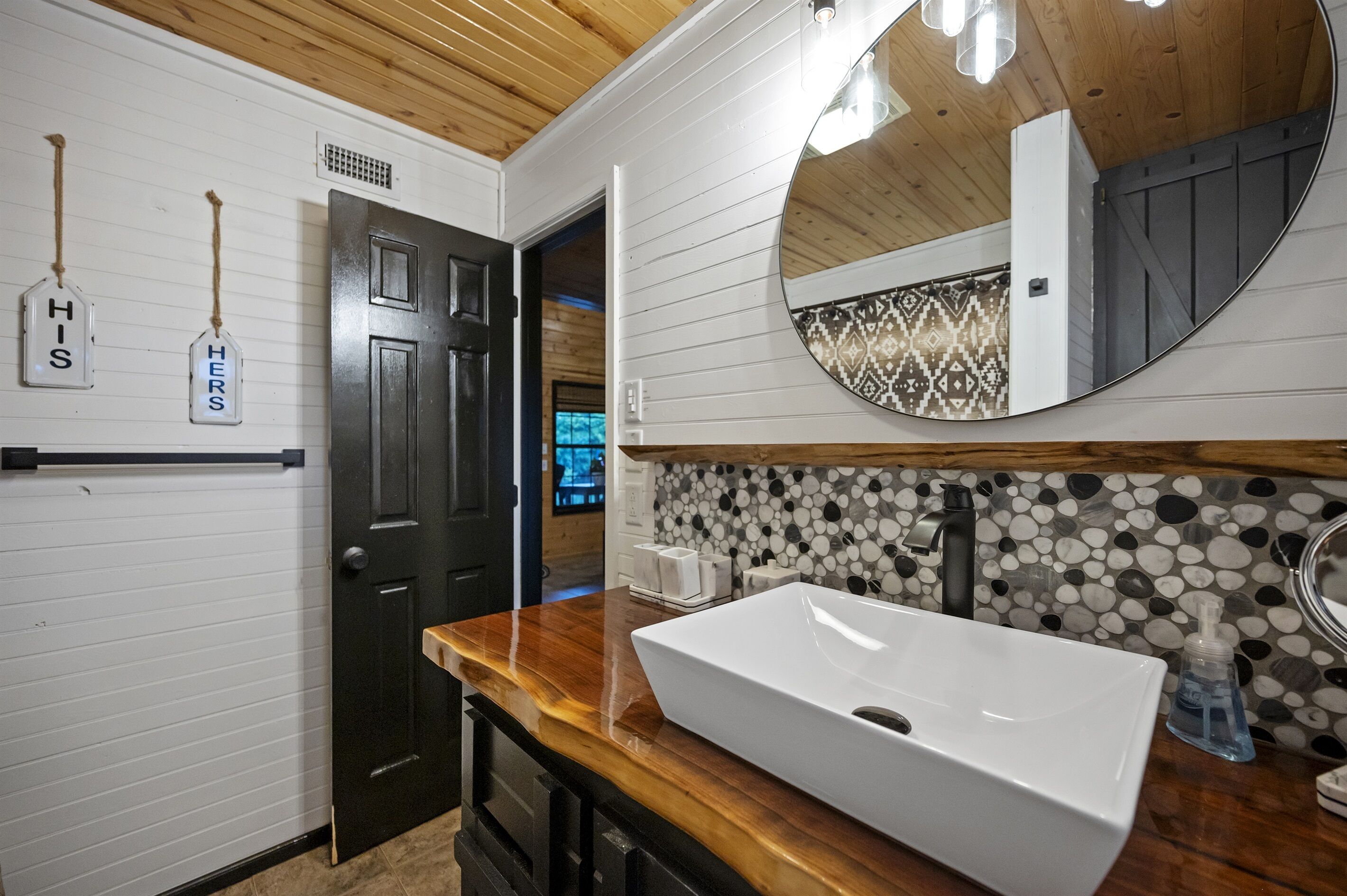 Un baño con un lavamanos blanco, una encimera de madera, una puerta negra y un espejo con un revestimiento de azulejos decorativos.