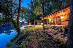 A cabin by a lake with a stone pathway leading to the entrance, surrounded by trees. - Cozy Cabin on Mt Fork River *riverfront* (Broken Bow)