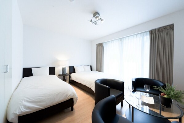 Quarto de hotel com duas camas, uma mesa pequena com uma cadeira e uma janela grande com cortinas.