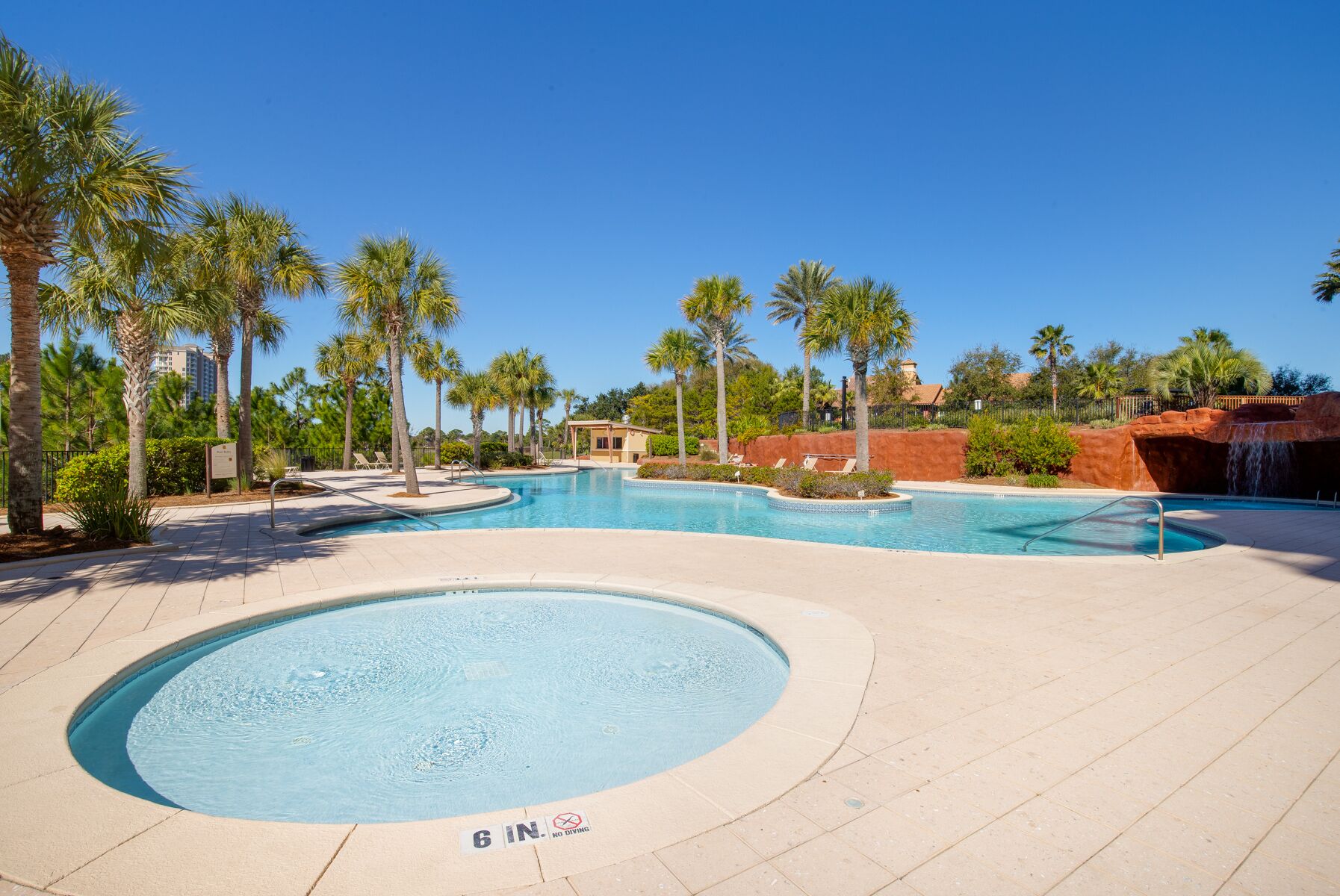 A large outdoor swimming pool with a 6 ft depth marker, surrounded by palm trees and a waterfall feature.