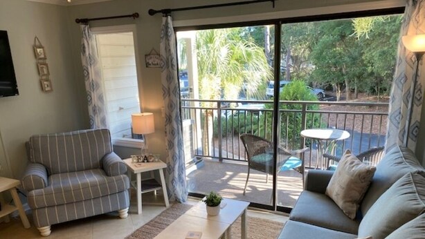A living room with a sofa, armchair, and a small table. There is a balcony with a view of trees and parked cars. - BEACHFRONT KING BED VILLA, PRIVATE BALCONY, SELF CHECKIN, FLEXIBLE CANCELLATION (Hilton Head Island)