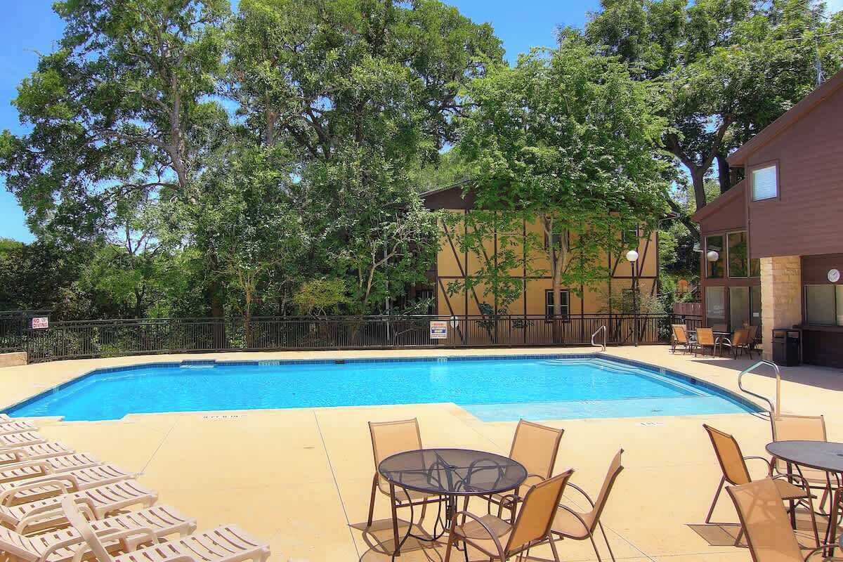 A swimming pool with lounge chairs and tables, surrounded by trees and a building with large windows.