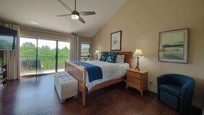 A bedroom with a bed, a ceiling fan, a bedside table with a lamp, a chair, a bookshelf, and a balcony with a view.