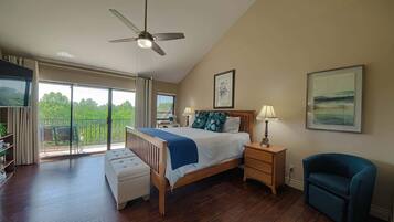 A bedroom with a bed, a ceiling fan, a bedside table with a lamp, a chair, a bookshelf, and a balcony with a view.