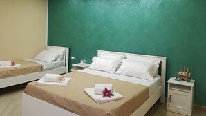 Une chambre d’hôtel avec deux lits, chacun agrémenté de draps blancs et d’une couvre-lit beige. On y trouve des tables de chevet blanches, sur lesquelles reposent de petites lampes, et une maquette de bateau sur l’une d’elles. Les murs sont peints en vert et le plafond est doté d’un éclairage encastré.