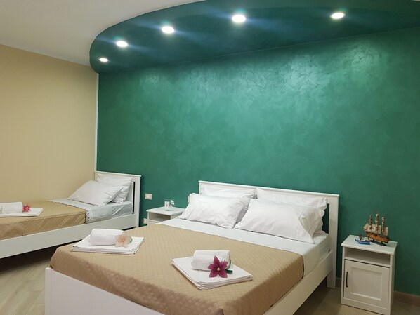 Une chambre d’hôtel avec deux lits, chacun agrémenté de draps blancs et d’une couvre-lit beige. On y trouve des tables de chevet blanches, sur lesquelles reposent de petites lampes, et une maquette de bateau sur l’une d’elles. Les murs sont peints en vert et le plafond est doté d’un éclairage encastré.